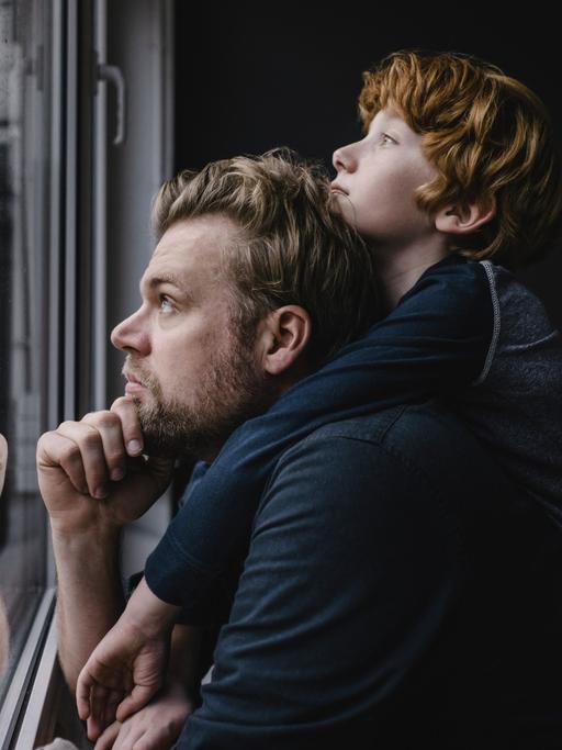Ein Vater, dessen Sohn über seinen Schultern lehnt, schaut aus dem Fenster.