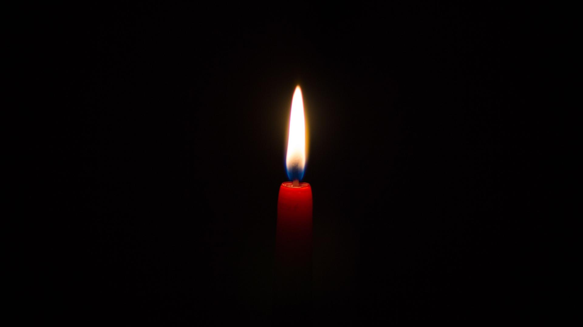 Vor einem schwarzen Hintergrund leuchtet eine Kerzenflamme. Die Kerze ist schmal und rot.