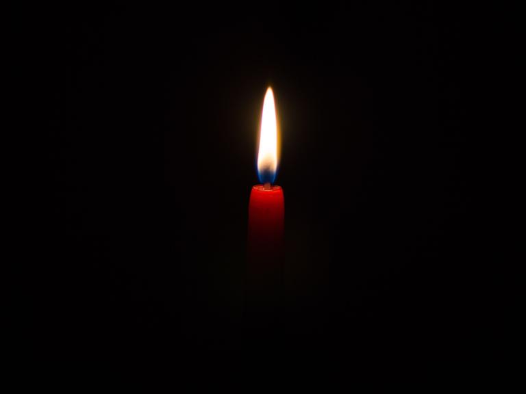 Vor einem schwarzen Hintergrund leuchtet eine Kerzenflamme. Die Kerze ist schmal und rot.