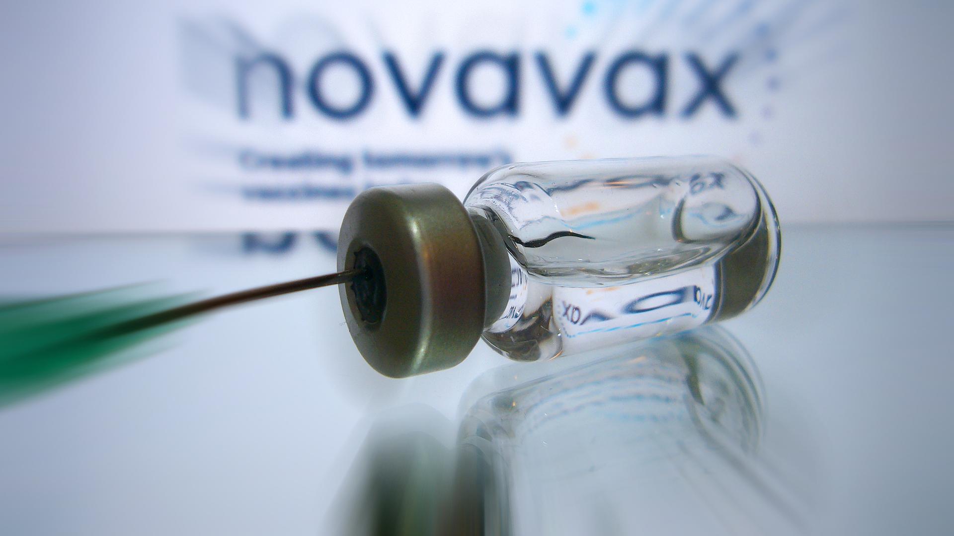 Eine Kanüle zieht eine Impfdosis aus einen Fläschchen auf. Im Hintergrund: das Logo von Novavax (Symbolfoto) Eine Kanüle zieht eine Impfdosis aus einen Fläschchen auf. Im Hintergrund: das Logo von Novavax (Symbolfoto)