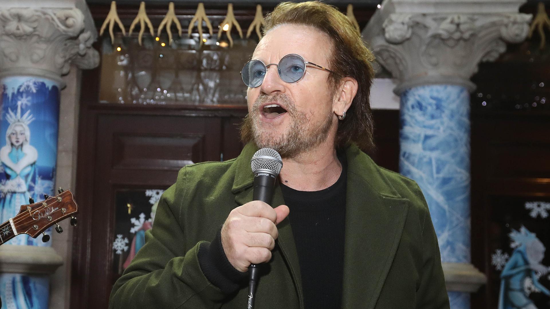 Bono singt in ein Mikrofon. Er hat einen Drei-Tage-Bart und trägt eine Brille mit bläulich getönten Gläsern. Bono singt in ein Mikrofon. Er hat einen Drei-Tage-Bart und trägt eine Brille mit bläulich getönten Gläsern.