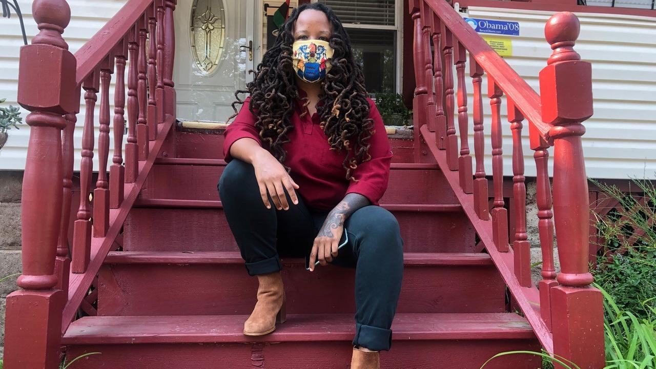 Imani Oakley sitzt auf der Treppe ihres Wohnhauses in der Stadt Montclair. Sie hat lange, schwarze Haare und trägt einen bunten Mund-Nasen-Schutz im Gesicht. Imani Oakley sitzt auf der Treppe ihres Wohnhauses in der Stadt Montclair. Sie hat lange, schwarze Haare und trägt einen bunten Mund-Nasen-Schutz im Gesicht.