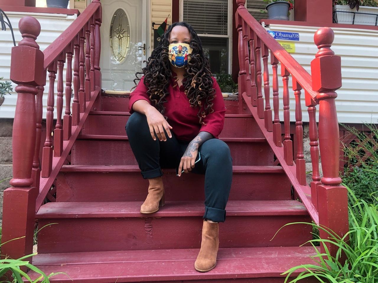 Imani Oakley sitzt auf der Treppe ihres Wohnhauses in der Stadt Montclair. Sie hat lange, schwarze Haare und trägt einen bunten Mund-Nasen-Schutz im Gesicht. Imani Oakley sitzt auf der Treppe ihres Wohnhauses in der Stadt Montclair. Sie hat lange, schwarze Haare und trägt einen bunten Mund-Nasen-Schutz im Gesicht.