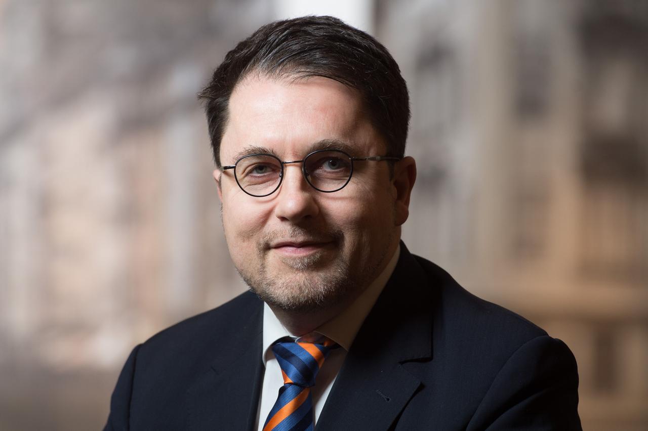 Ein Mann in Anzug und Krawatte, mit Seitenscheitel, Brille und Drei-Tage-Bart blickt in die Kamera. Ein Mann in Anzug und Krawatte, mit Seitenscheitel, Brille und Drei-Tage-Bart blickt in die Kamera.