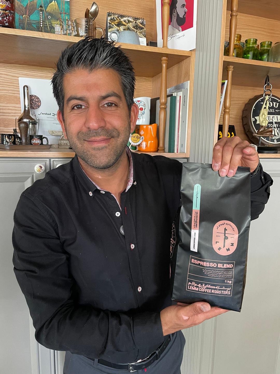 Vor einem Regal steht ein Mann mit schwarzen Haaren und schwarzem Hemd und hebt eine Kaffeepackung hoch. Vor einem Regal steht ein Mann mit schwarzen Haaren und schwarzem Hemd und hebt eine Kaffeepackung hoch.