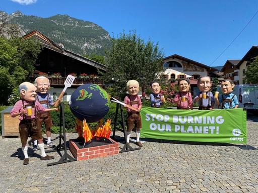 Aktivisten von Oxfam protestieren mit Politiker-Köpfen am Rande des G7-Gipfels auf Schloss Elmau gegen die Klimapolitik der Industrienationen. Die Figuren Scholz. Biden und Johnson drehen dabei eine Weltkugel aus Pappmaché über einer Feuerstelle aus Pappe.