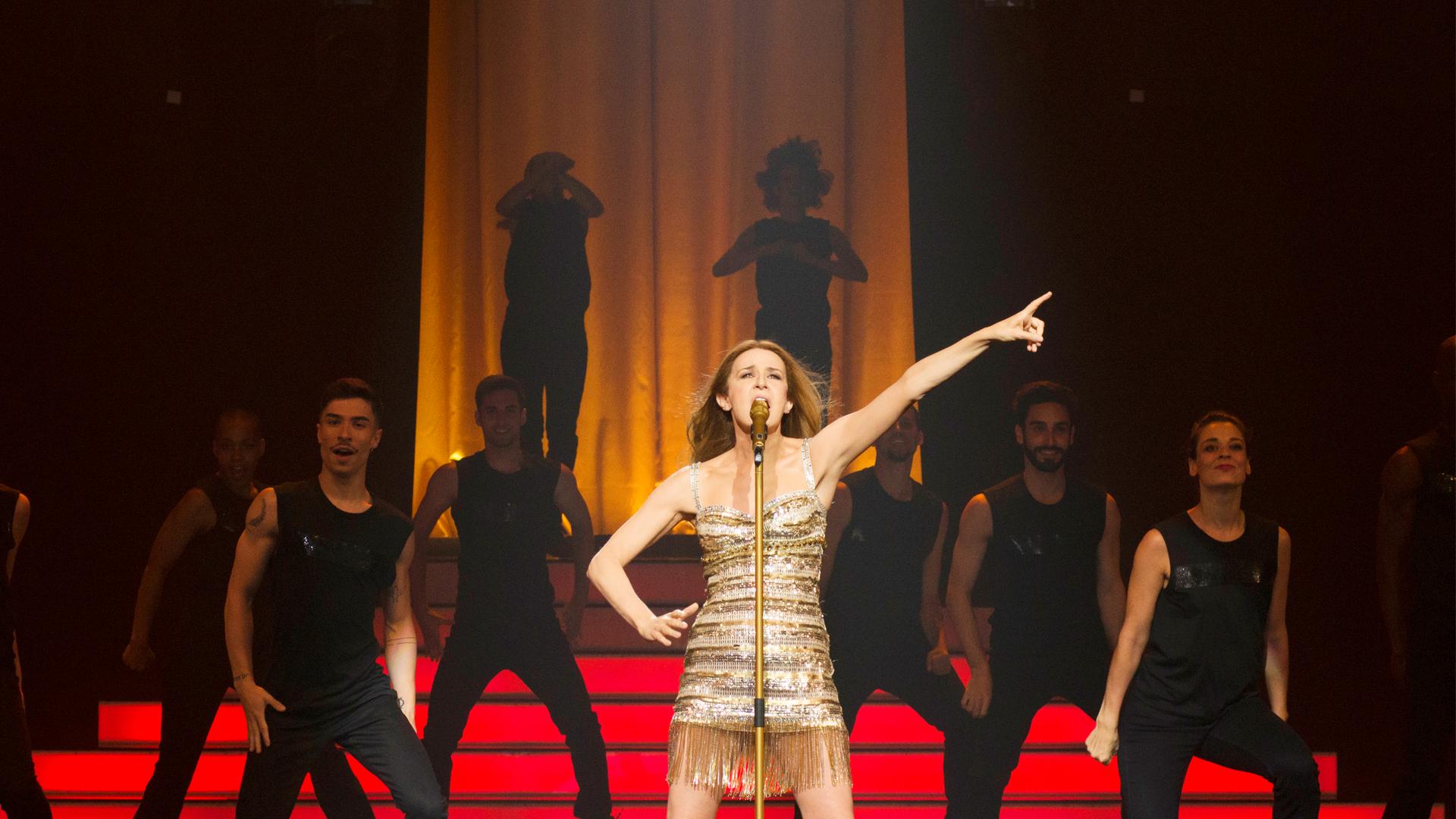 Die Darstellerin der "Aline" in gewohnter Céline-Dion-Pose auf der großen Bühne, im Hintergrund Tänzer*innen vor roten Stufen.