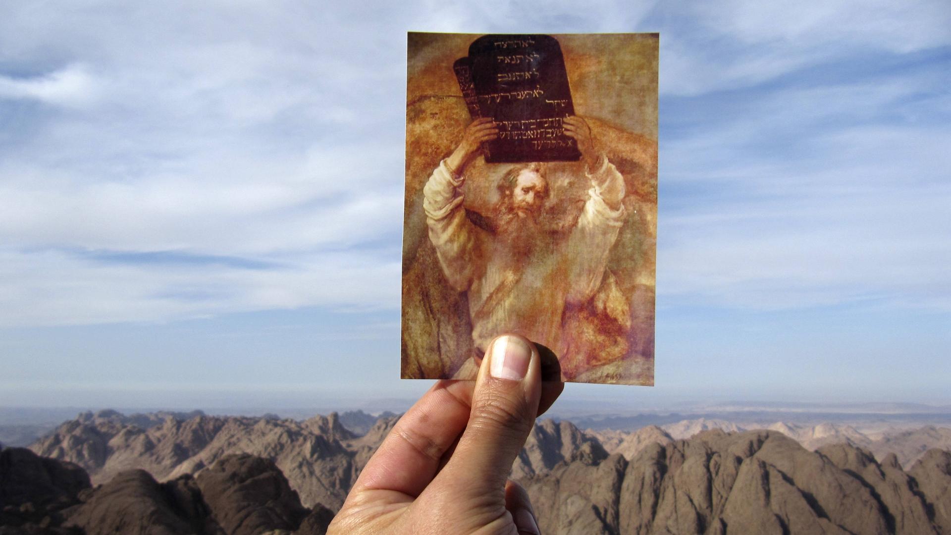 Eine Hand hält vor einem Gebirge eine Postkarte hoch, auf der ein Rubens Gemälde zu sehen ist, das Moses mit der Tafel der zehn Gebote zeigt. Eine Hand hält vor einem Gebirge eine Postkarte hoch, auf der ein Rubens Gemälde zu sehen ist, das Moses mit der Tafel der zehn Gebote zeigt.