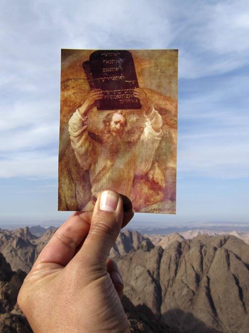 Eine Hand hält vor einem Gebirge eine Postkarte hoch, auf der ein Rubens Gemälde zu sehen ist, das Moses mit der Tafel der zehn Gebote zeigt.