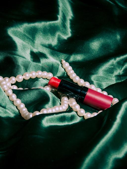 Ein Lippenstift und eine Perlenkette liegen auf einem grünen Tuch.