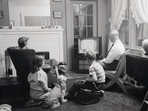 Schwarzweißfoto einer Familie mit zwei Kindern in den 1950er Jahren, die gemeinsam mit einem Hund vor dem Fernseher sitzt.