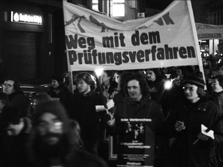 Historische schwarz-weiß-Aufnahme von einem Fackelmarsch gegen die Pruefung zur Kriegsdienstverweigerung in Bonn im Dezember 1977. Auf einem Transparent steht "Weg mit dem Prüfungsverfahren".