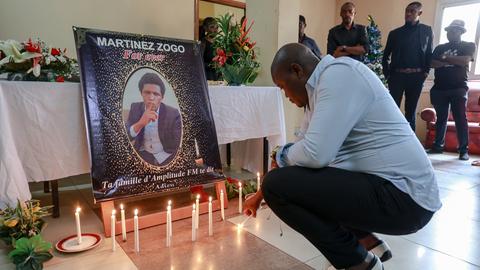 Ein Man zündet vor einem Bild eine Kerze an. Das Bild zeigt den getöteten Journalisten Martinez Zogo.