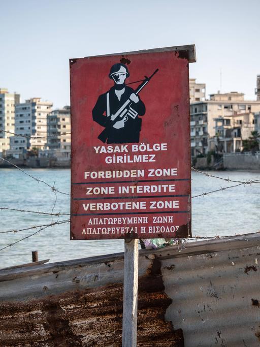 Ein rotes Warnschild zeigt, dass der dahinter liegende Strand militätisches Sperrgebiet ist. Im Hintergrund sieht man verfallene Hotelgebäude.