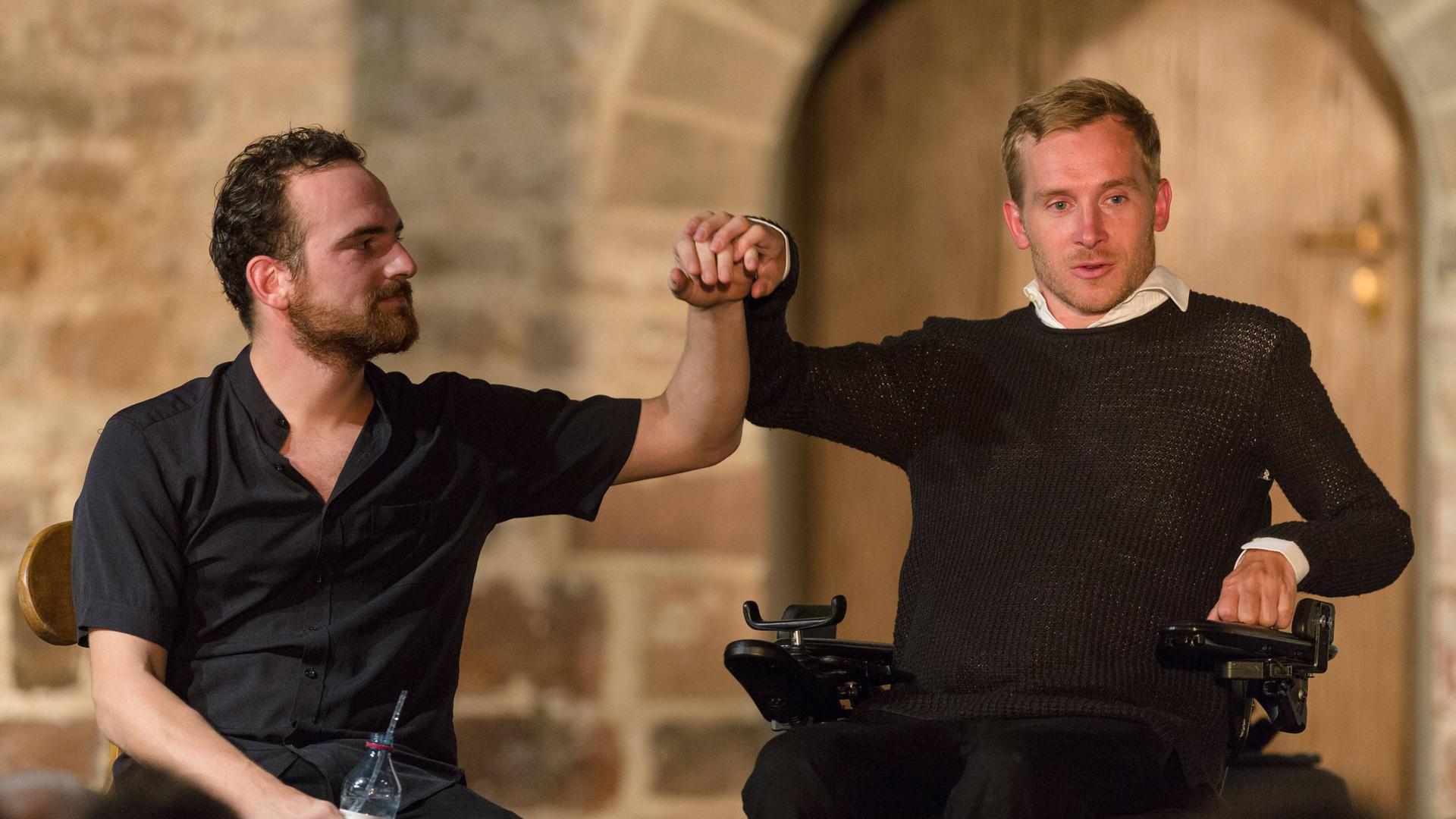 Rechts auf einer Bühne sitzt ein Mann im Rollstuhl und legt seine Hand auf die erhobene Hand eines Mannes, der links von ihm auf einem Stuhl sitzt. Rechts auf einer Bühne sitzt ein Mann im Rollstuhl und legt seine Hand auf die erhobene Hand eines Mannes, der links von ihm auf einem Stuhl sitzt.