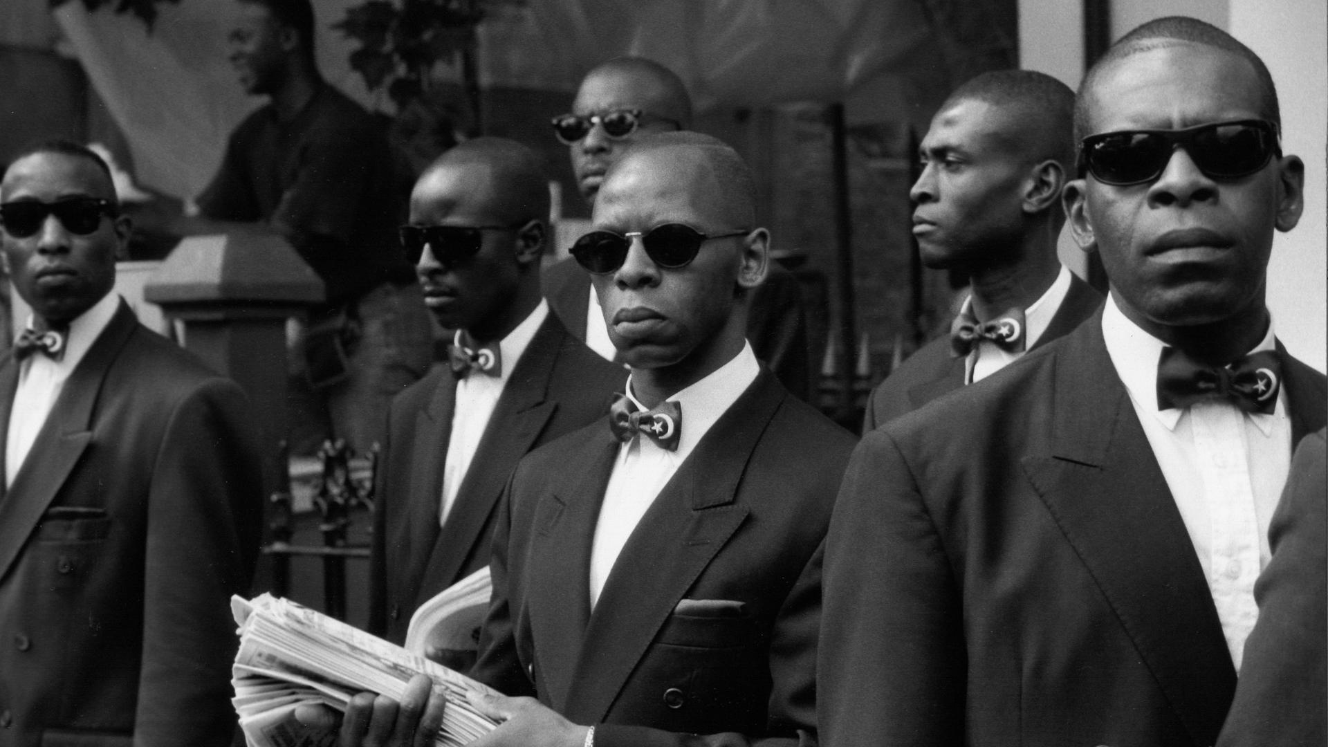 Mitglieder der 'Nation of Islam' stehen in ihren Anzügen und mit schwarzen Sonnenbrillen in Notting Hill auf der Straße mit ihrer Mitgliederzeitschrift 1991. Mitglieder der 'Nation of Islam' stehen in ihren Anzügen und mit schwarzen Sonnenbrillen in Notting Hill auf der Straße mit ihrer Mitgliederzeitschrift 1991.