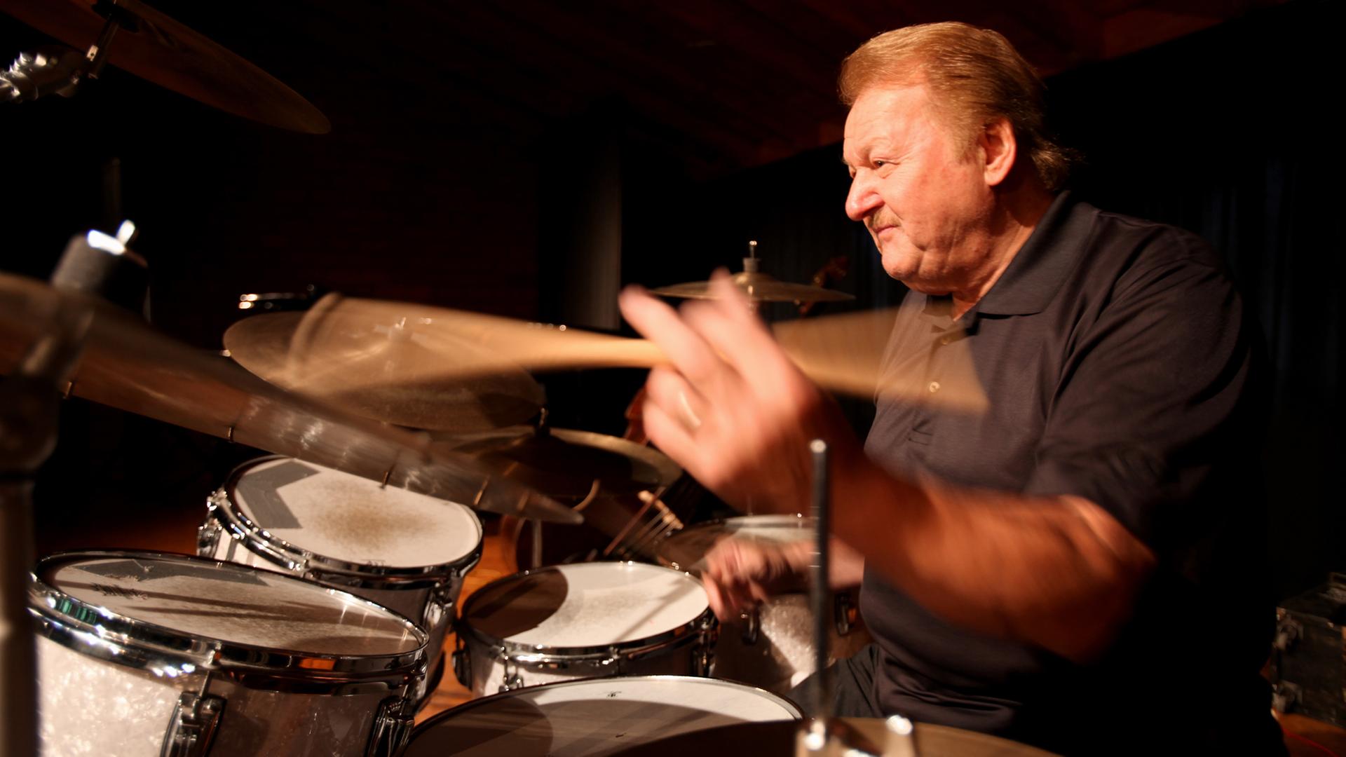 Ein älterer blonde Herr sitzt am Drumset und betätigt mit schnellen Bewegungen eines Drumsticks das Becken. Ein älterer blonde Herr sitzt am Drumset und betätigt mit schnellen Bewegungen eines Drumsticks das Becken.