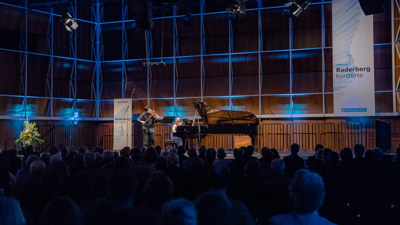 Ein Geiger konzertiert mit einer Pianistin vor Publikum auf einem Podium. Der Konzertsaal ist holzvertäfelt und schimmert in bläulichem Licht. An den Wänden hängen Banner mit der Aufschrift "Raderbergkonzerte Deutschlandfunk".