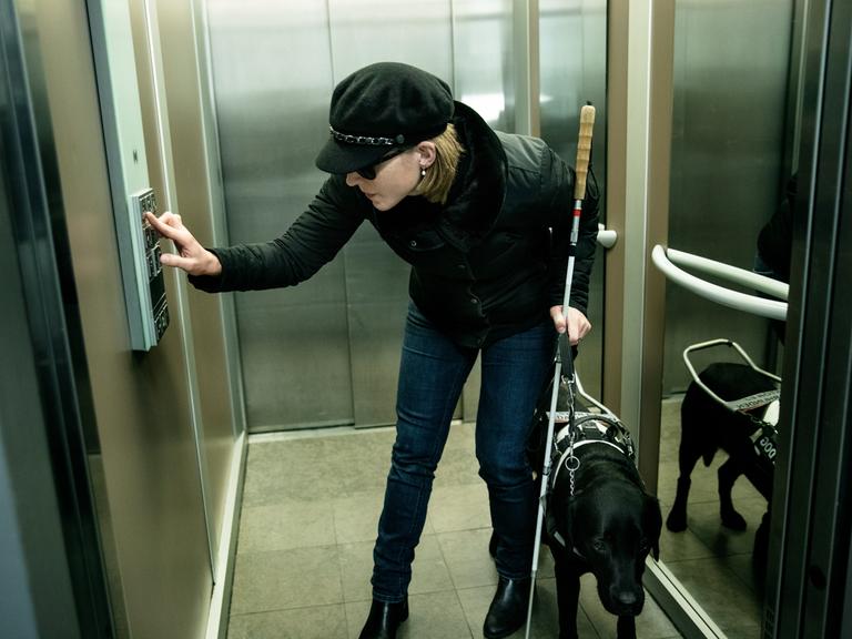 Eine blinde Frau mit Blindenhund an der Leine drückt in einem Fahrstuhl einen Knopf.
