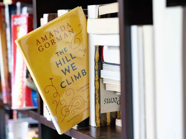 Ein gelbes Buch mit dem englischen  Titel "The hill we climb" scheint fast aus einem Bücherregal herauszufallen