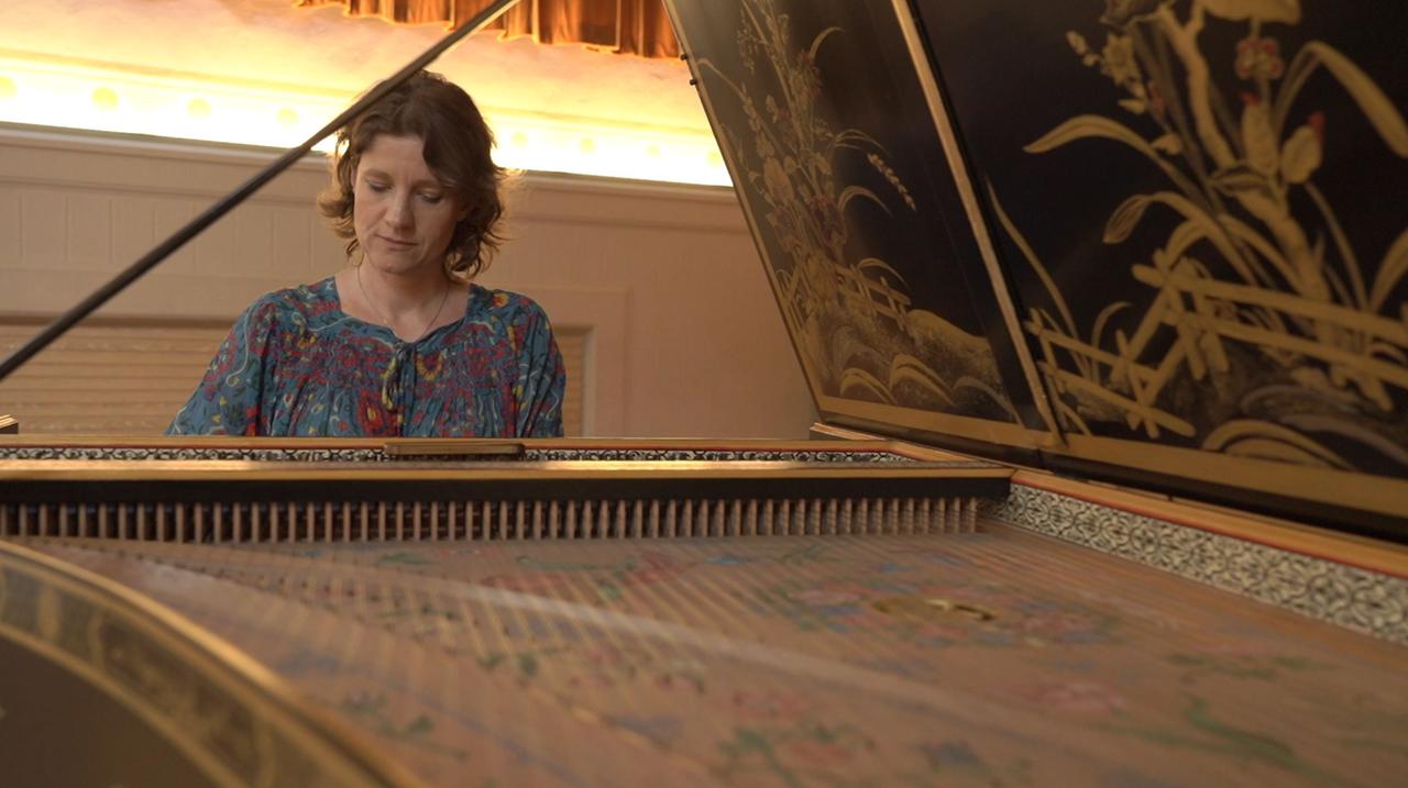 Eine Frau mit schulterlangen Haaren sitzt an einem aufgeklappten, historischen Tasteninstrument und spielt. Eine Frau mit schulterlangen Haaren sitzt an einem aufgeklappten, historischen Tasteninstrument und spielt.