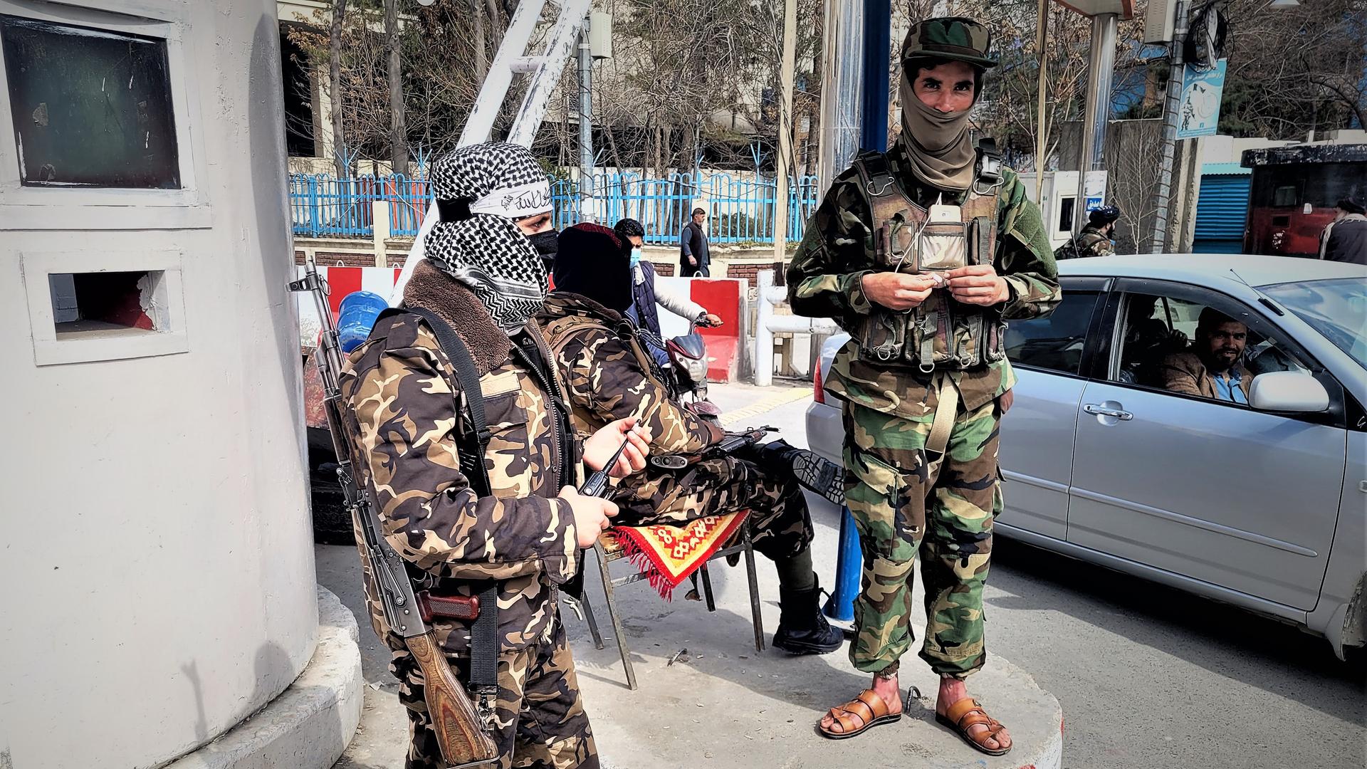 Zwei Taliban in grüner und brauner Tarnuniform stehen bewaffnet an einem Checkpoint in Kabul. Im Hintergrund sitzt ein weiterer Taliban auf einem Stuhl und schaut zu einem Autofahrer. Zwei Taliban in grüner und brauner Tarnuniform stehen bewaffnet an einem Checkpoint in Kabul. Im Hintergrund sitzt ein weiterer Taliban auf einem Stuhl und schaut zu einem Autofahrer.