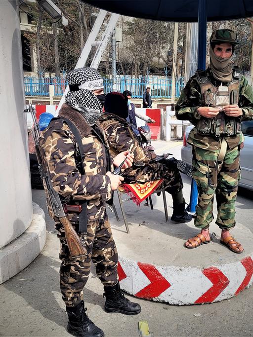 Zwei Taliban in grüner und brauner Tarnuniform stehen bewaffnet an einem Checkpoint in Kabul. Im Hintergrund sitzt ein weiterer Taliban auf einem Stuhl und schaut zu einem Autofahrer.