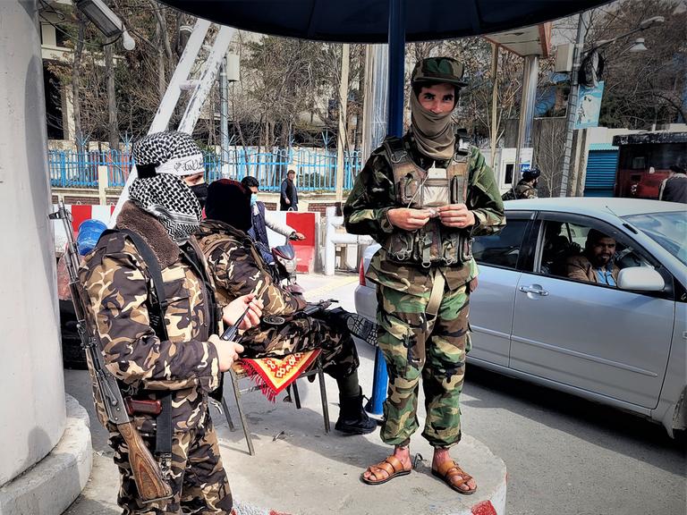 Zwei Taliban in grüner und brauner Tarnuniform stehen bewaffnet an einem Checkpoint in Kabul. Im Hintergrund sitzt ein weiterer Taliban auf einem Stuhl und schaut zu einem Autofahrer.