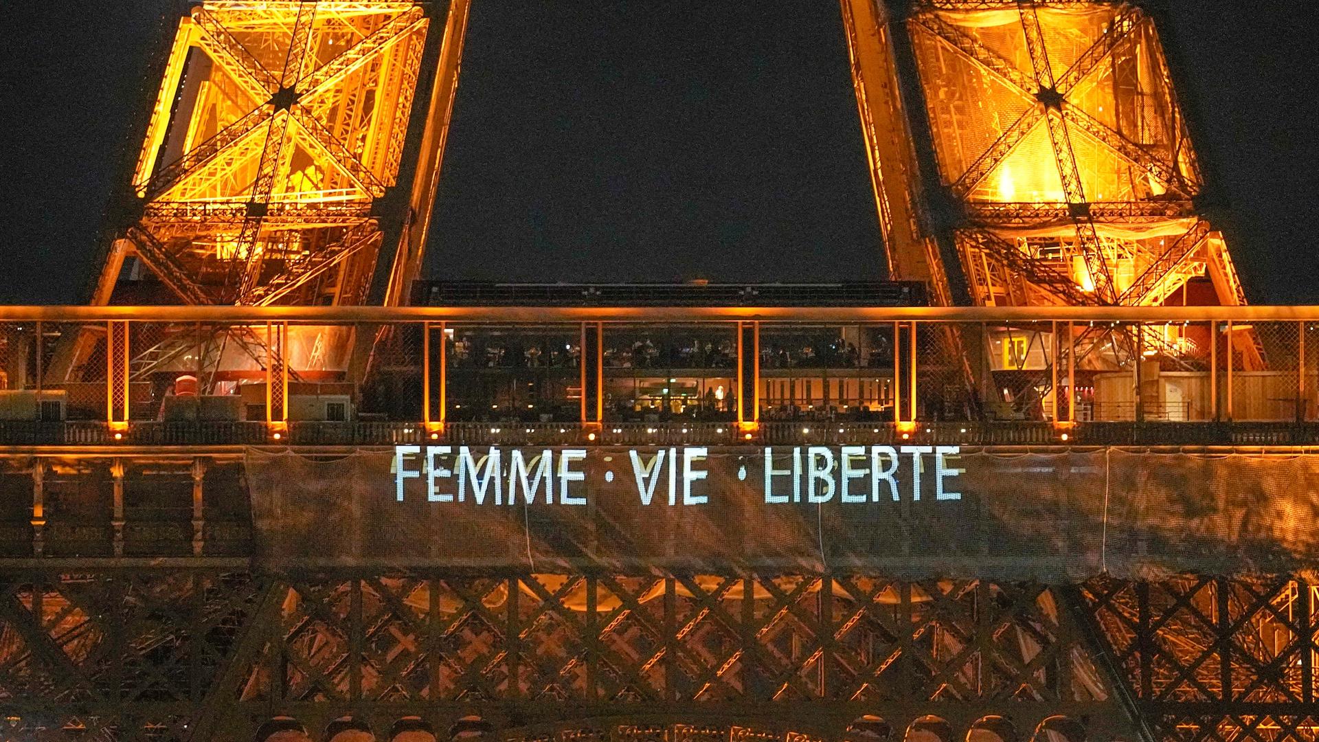 Der Eiffel-Turm leuchtet in der Nacht. Man sieht  den Spruch:  "Femme, Vie, Liberté". Das bedeutet "Frau, Leben, Freiheit".