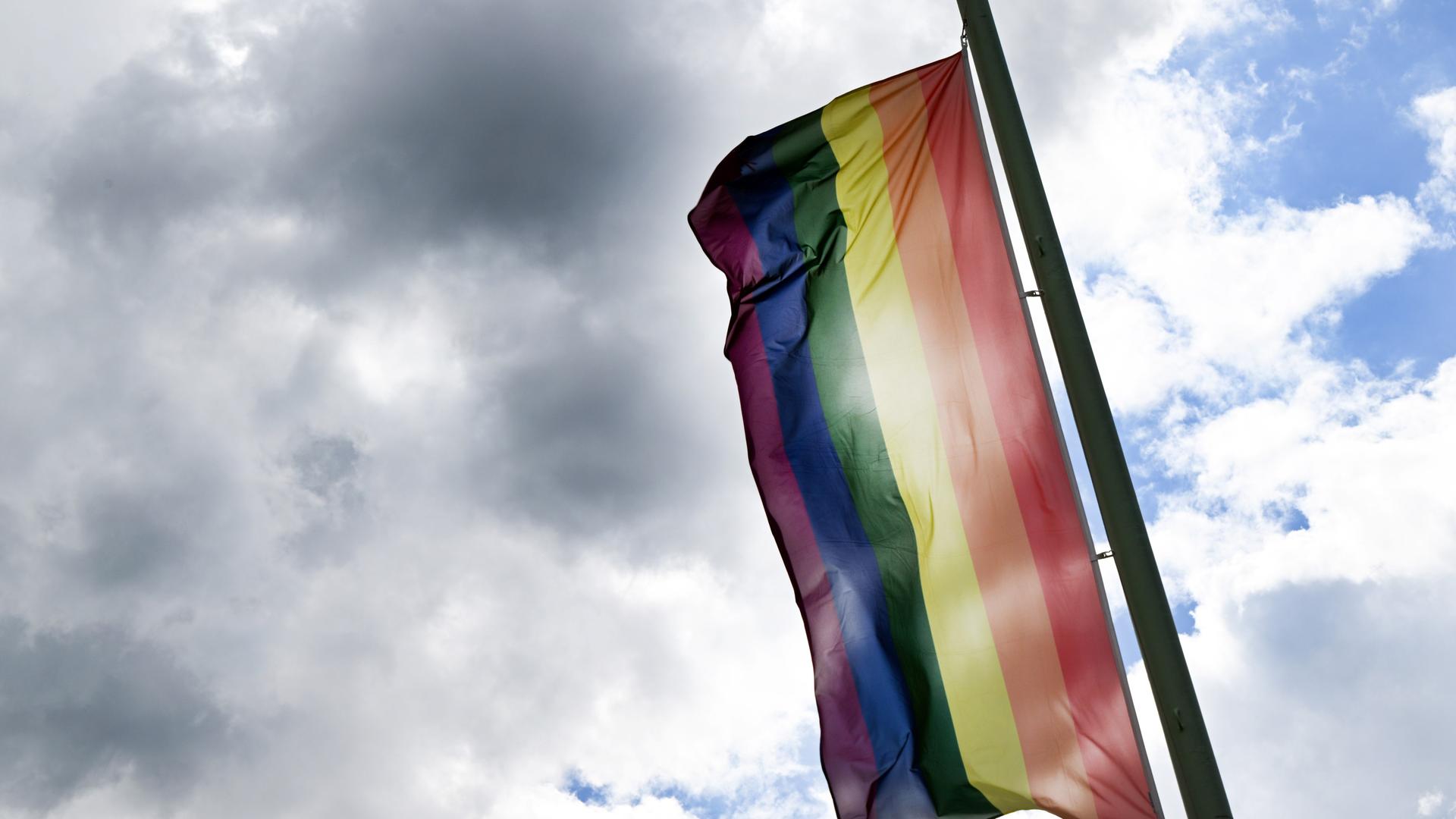 Eine Regenbogenflagge weht an einem Fahnenmast. Der Himmel ist bedeckt, auch mit grauen Wolken. Eine Regenbogenflagge weht an einem Fahnenmast. Der Himmel ist bedeckt, auch mit grauen Wolken.