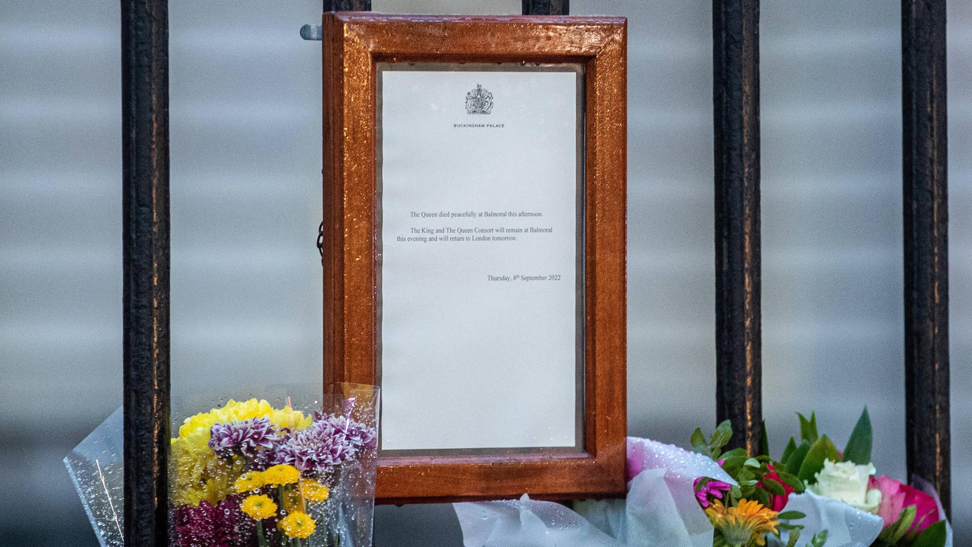 Ein Blatt Papier mit der Todesanzeige von Königin Elisabeth hängt in einem braunen Rahmen an einem Zaun, daneben stehen Blumen. Ein Blatt Papier mit der Todesanzeige von Königin Elisabeth hängt in einem braunen Rahmen an einem Zaun, daneben stehen Blumen.