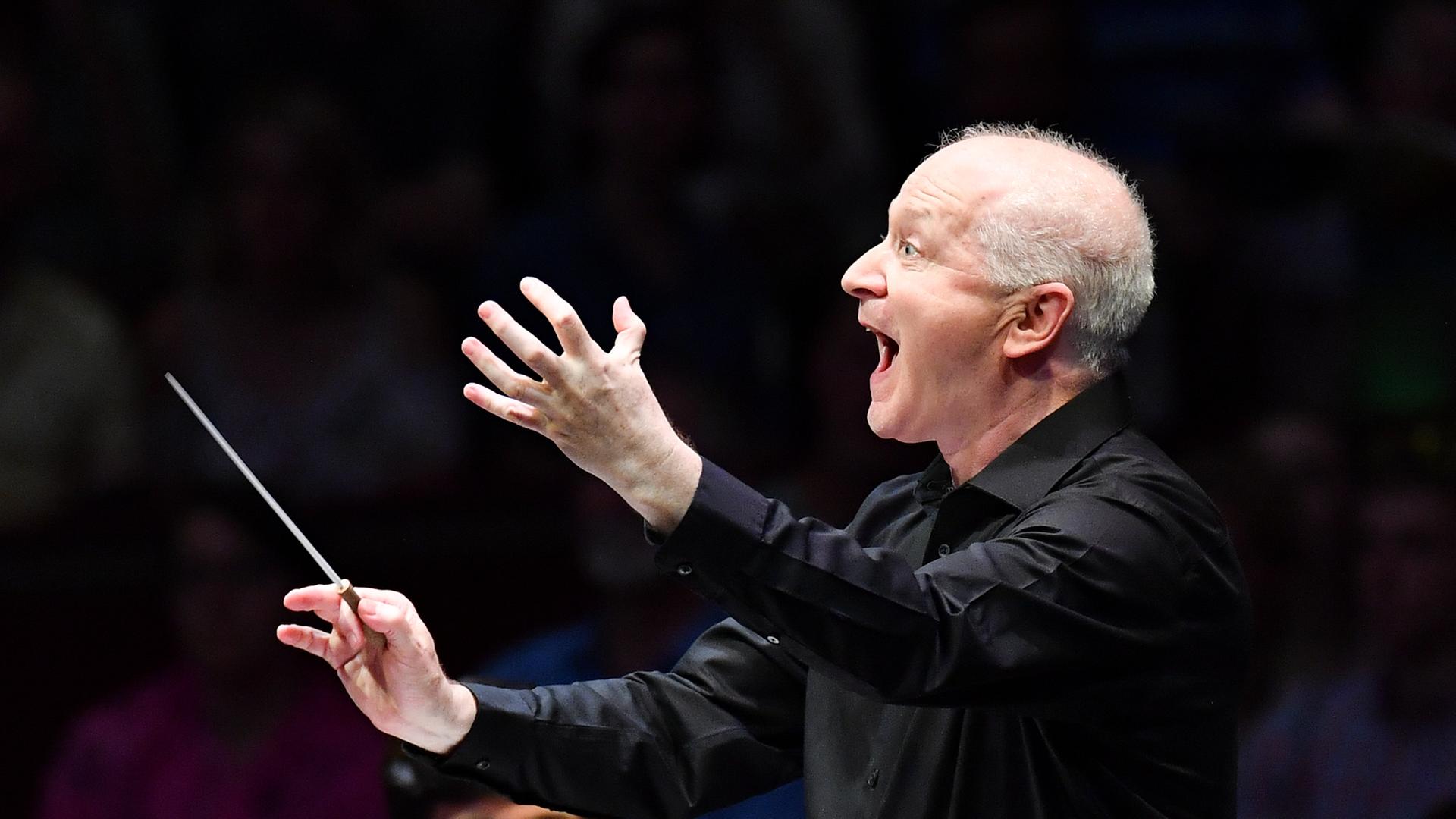 Der Dirigent Sir George Benjamin feuert mit wilder Geste ein Orchester an. Der Dirigent Sir George Benjamin feuert mit wilder Geste ein Orchester an.