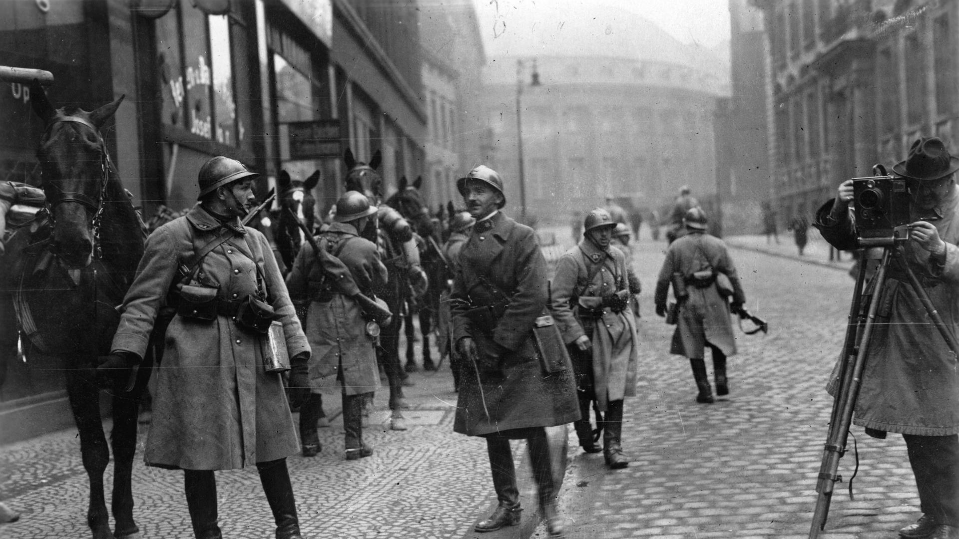 Französische Soldaten in Essen bei der Besetzung des Ruhrgebiets durch französische Truppen. Links hält ein Soldat ein Pferd, andere Soldaten gehen in der Straße umher, ganz rechts steht ein Fotograf mit seiner aufgebauten Kamera.