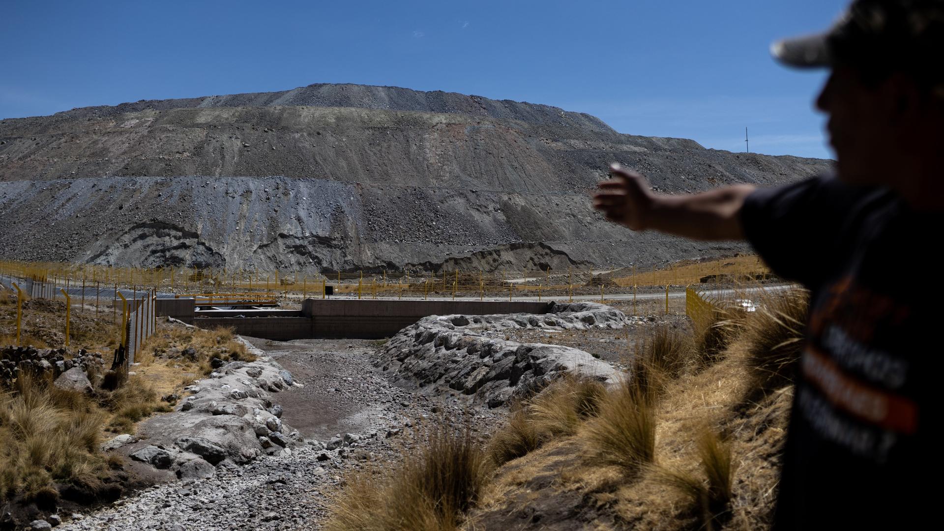 Vidal Merma steht nahe einer Kupfermine in Espinar. Nur sein Schatten ist erkennbar. Er zeigt auf Abfälle. Im Hintergrund sind Zäune und ein grauer Berg aus Steinen und Sand zu sehen. Vidal Merma steht nahe einer Kupfermine in Espinar. Nur sein Schatten ist erkennbar. Er zeigt auf Abfälle. Im Hintergrund sind Zäune und ein grauer Berg aus Steinen und Sand zu sehen.