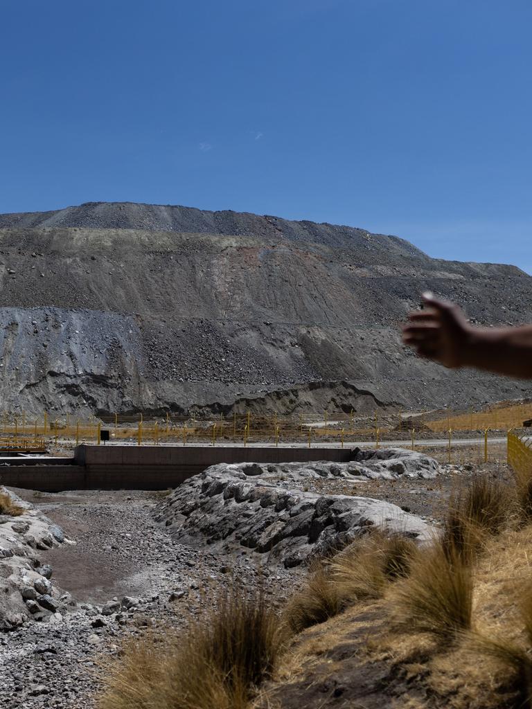 Vidal Merma steht nahe einer Kupfermine in Espinar. Nur sein Schatten ist erkennbar. Er zeigt auf Abfälle. Im Hintergrund sind Zäune und ein grauer Berg aus Steinen und Sand zu sehen.