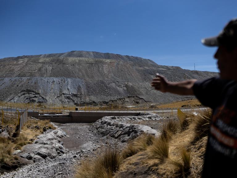 Vidal Merma steht nahe einer Kupfermine in Espinar. Nur sein Schatten ist erkennbar. Er zeigt auf Abfälle. Im Hintergrund sind Zäune und ein grauer Berg aus Steinen und Sand zu sehen.
