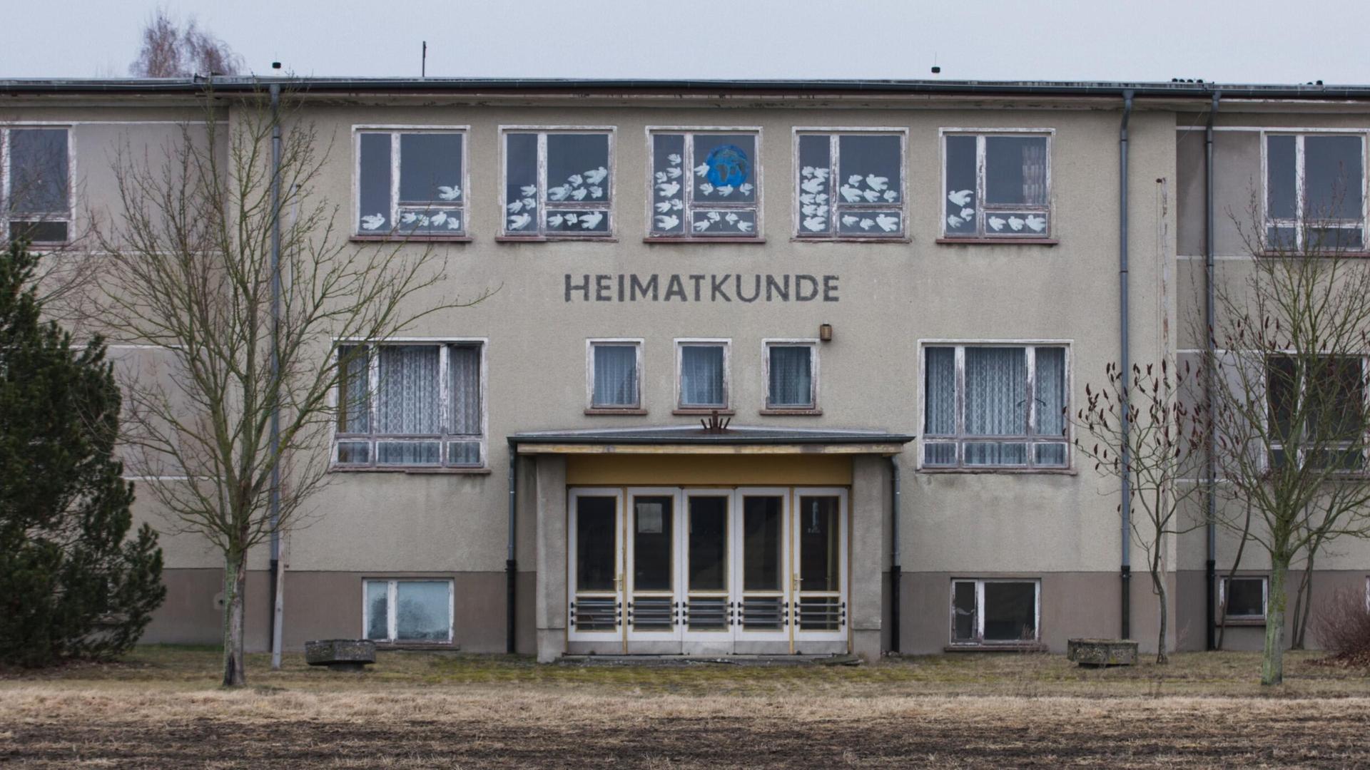 Ein großes, graues Gebäude auf einem winterlichen Feld. Es handelt sich um die Polytechnische Oberschule Bärenklau. Das Foto ist die Titeleinstellung des Films und zeigt die Front mit Eingangstür. Darüber steht der Schriftzug "Heimatkunde". Ein großes, graues Gebäude auf einem winterlichen Feld. Es handelt sich um die Polytechnische Oberschule Bärenklau. Das Foto ist die Titeleinstellung des Films und zeigt die Front mit Eingangstür. Darüber steht der Schriftzug "Heimatkunde".