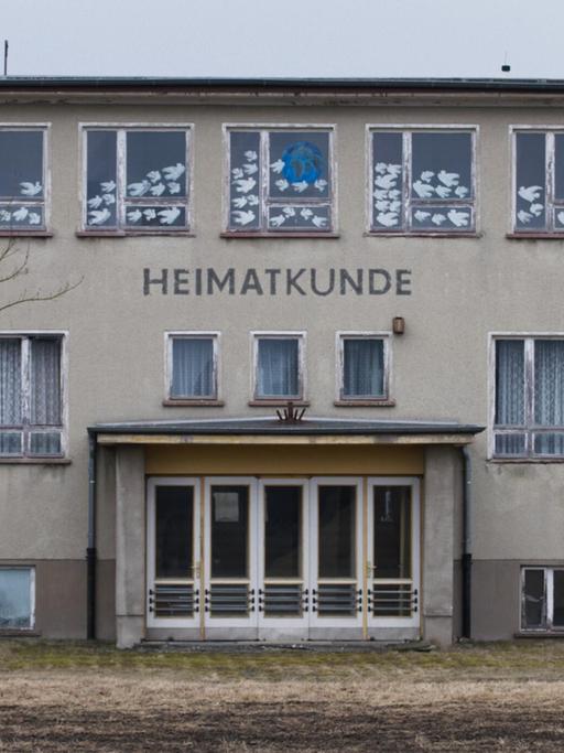 Ein großes, graues Gebäude auf einem winterlichen Feld. Es handelt sich um die Polytechnische Oberschule Bärenklau. Das Foto ist die Titeleinstellung des Films und zeigt die Front mit Eingangstür. Darüber steht der Schriftzug "Heimatkunde".
