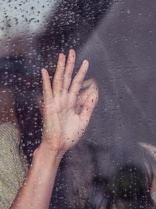 Eine trauernder Frau steht an einer verregneten Scheibe, an die sie ihre Hand drückt