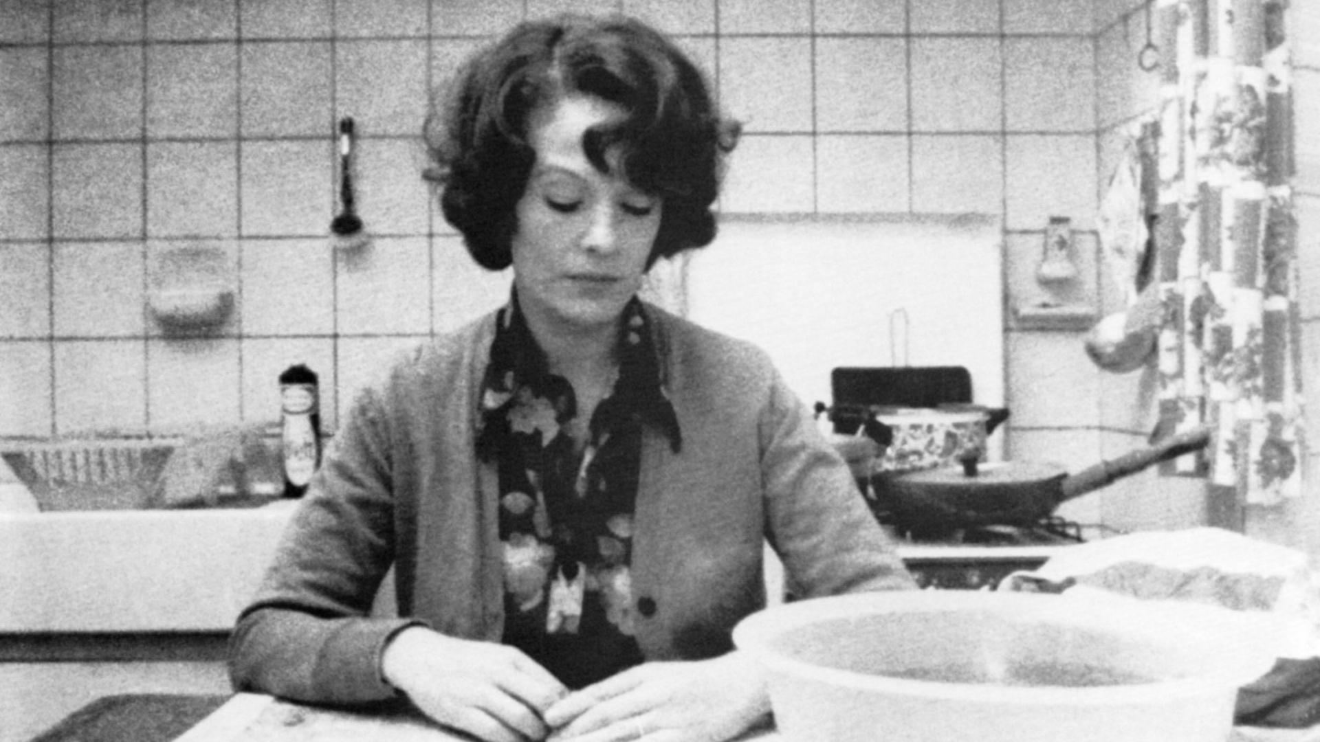 Auf dem Schwarz-Weiß-Bild sitzt eine Frau am Küchentisch. Vor ihr eine Schüssel mit Wasser und ein Sparschäler. Sie blickt nach unten, sie wirkt in sich gekehrt. 