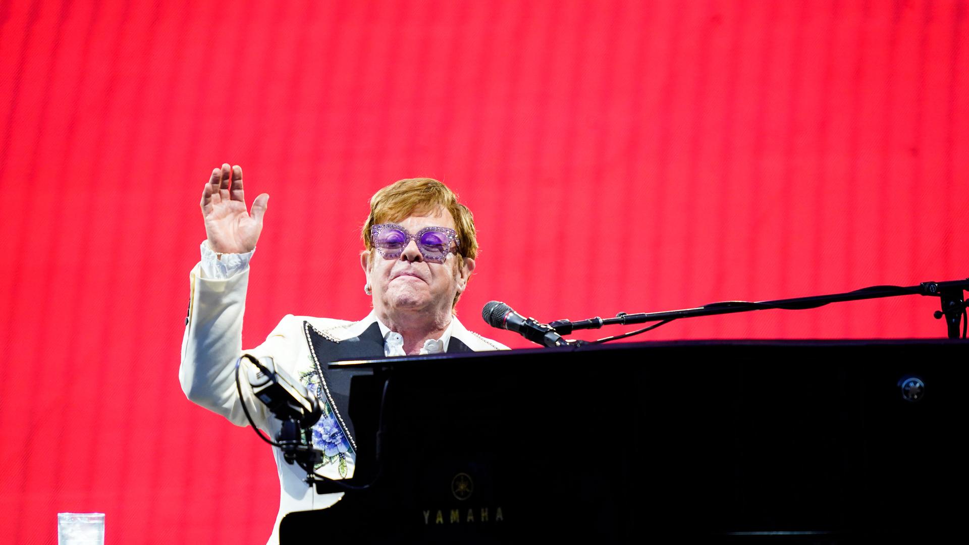 Der Musiker Elton John sitzt am Flügel auf einer Bühne vor einem roten Hintergrund