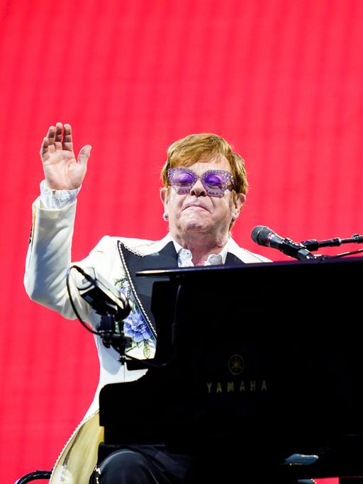 Der Musiker Elton John sitzt am Flügel auf einer Bühne vor einem roten Hintergrund