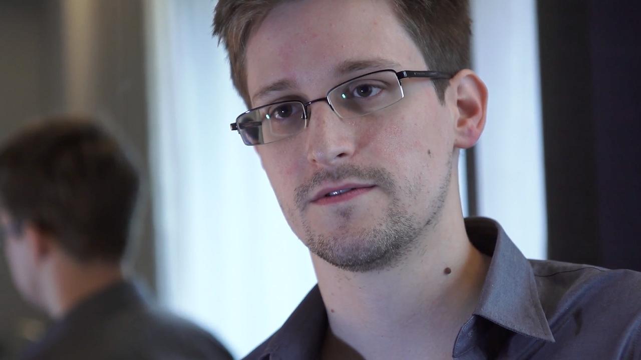 Edward Snowden mit grauem Hemd und Brille sitzt mit leicht schrägem Kopf so, dass sein Kopf sich im hinter ihm stehenden Spiegel spiegelt.