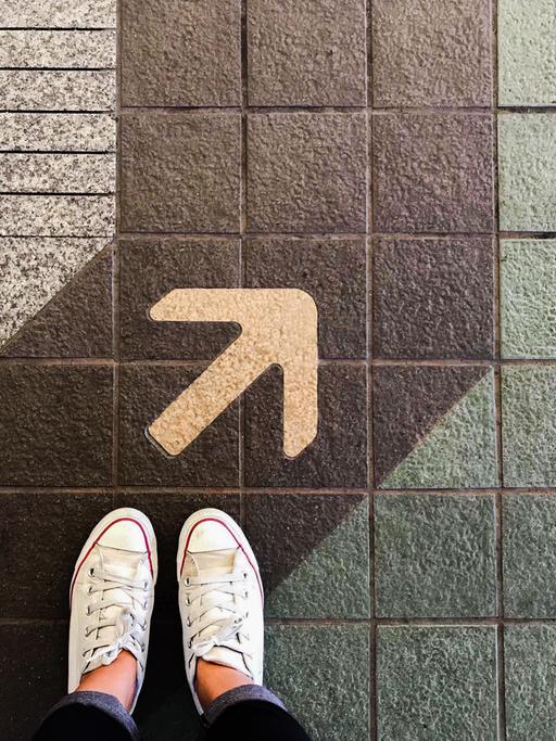 Die Füße einer Frau mit Turnschuhen stehen auf einer Straße, auf der eine Spur und ein Pfeil aufgemalt sind.
