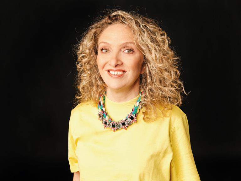 Katja Garmasch trägt ein gelbes Oberteil mit einer bunten Kette und steht vor einem schwarzen Hintergrund
