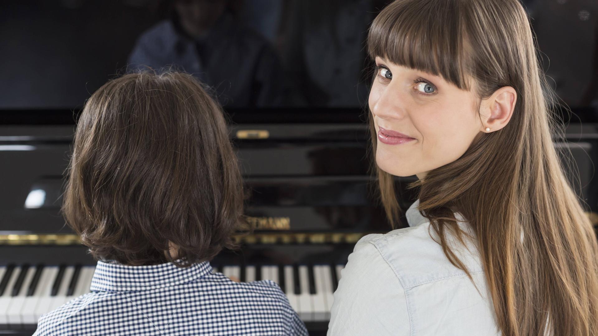 Eine Frau sitzt mit ihrem Kind am Klavier, das spielt, während sie über die Schulter in die Kamera schaut. Eine Frau sitzt mit ihrem Kind am Klavier, das spielt, während sie über die Schulter in die Kamera schaut.