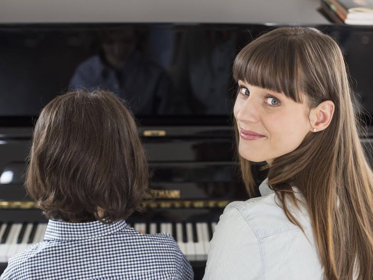 Eine Frau sitzt mit ihrem Kind am Klavier, das spielt, während sie über die Schulter in die Kamera schaut.