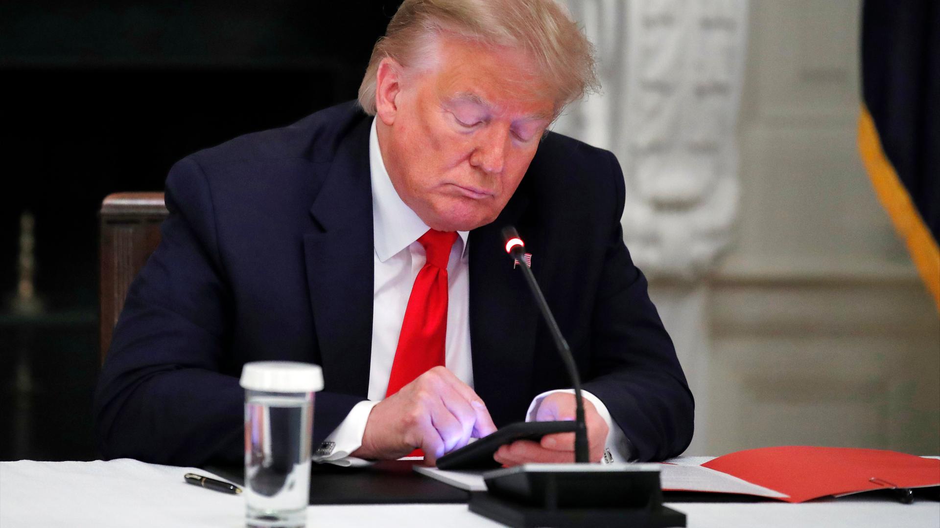 Donald Trump sitz mit gesenktem Blick an einem Konferenztisch und schaut auf das erleuchtete Display seines Smartphones.