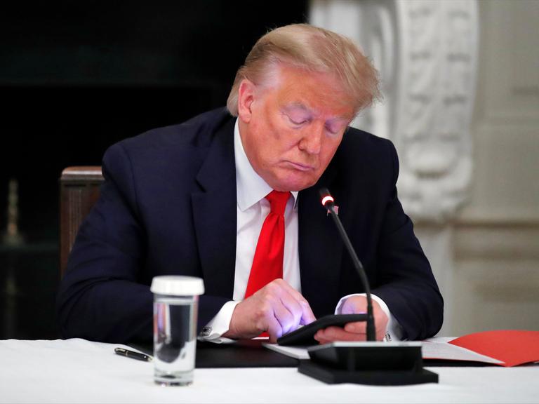 Donald Trump sitz mit gesenktem Blick an einem Konferenztisch und schaut auf das erleuchtete Display seines Smartphones.