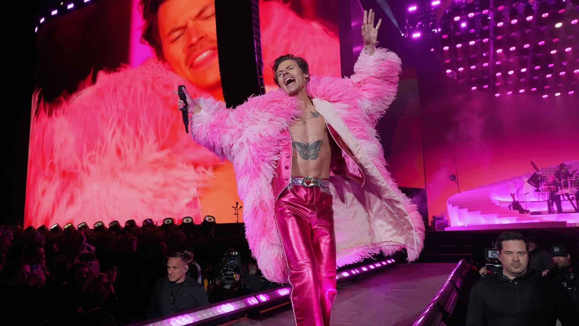 Harry Styles performt auf der Bühne. Er trägt einen Mantel aus pinkfarbenen Federn.